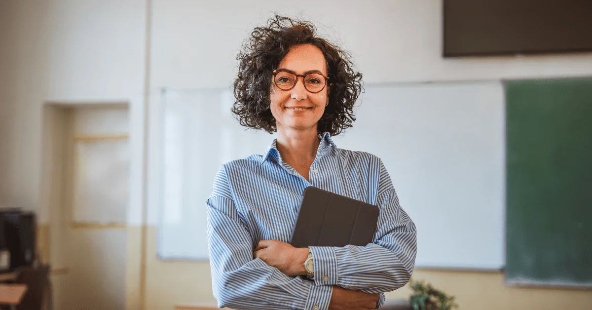 professora sorridente, de cabelos cacheados e óculos, vestindo uma camisa listrada azul e branca.