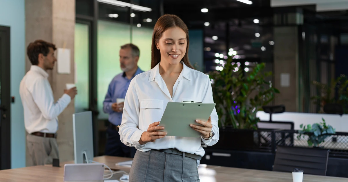 advogada sorridente em primeiro plano segurando uma prancheta, enquanto outros dois advogados conversam ao fundo em um escritório corporativo moderno.