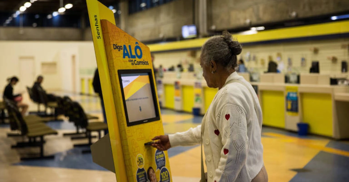 Mulher idosa retirando senha em totem de atendimento auto,ático de uma agência dos Correios. O concurso está previsto.